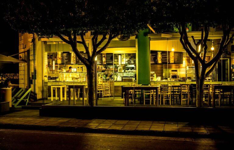 Exterior of a warmly lit urban café at night, featuring empty outdoor seating and trees lining the pavement.